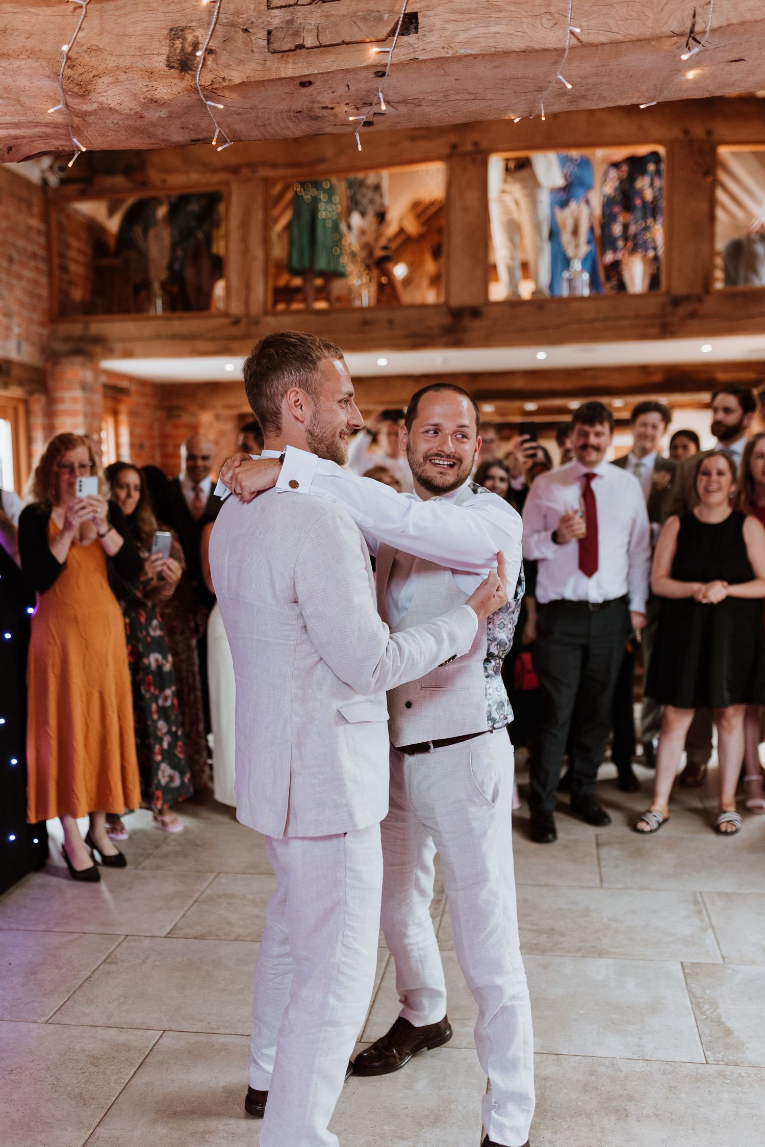 Two grooms share their first dance surrounded by cheering guests in a warmly lit Moat Hall Barn