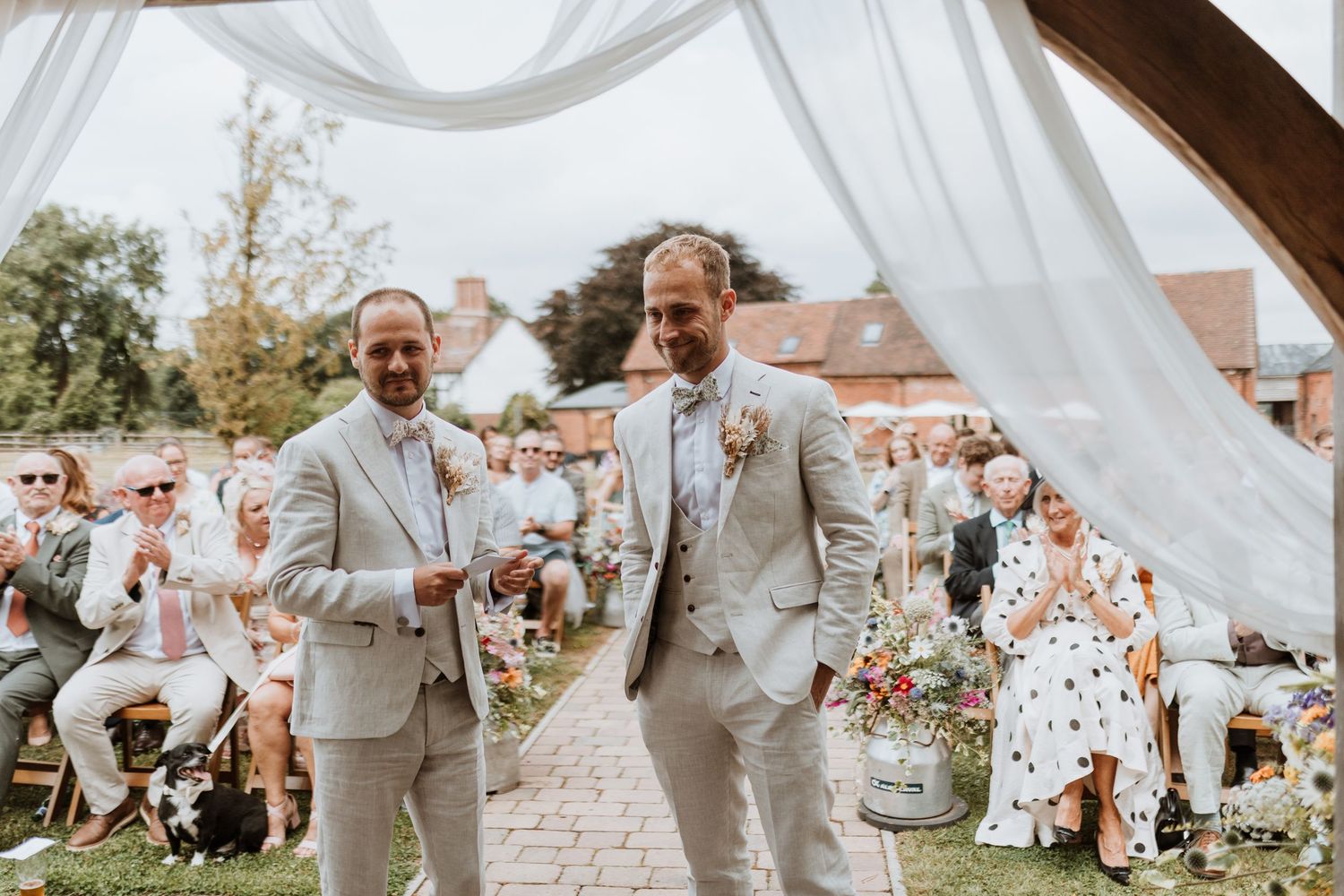 Two grooms in beige suits walk down the aisle at an outdoor wedding ceremony with guests seated on both sides.