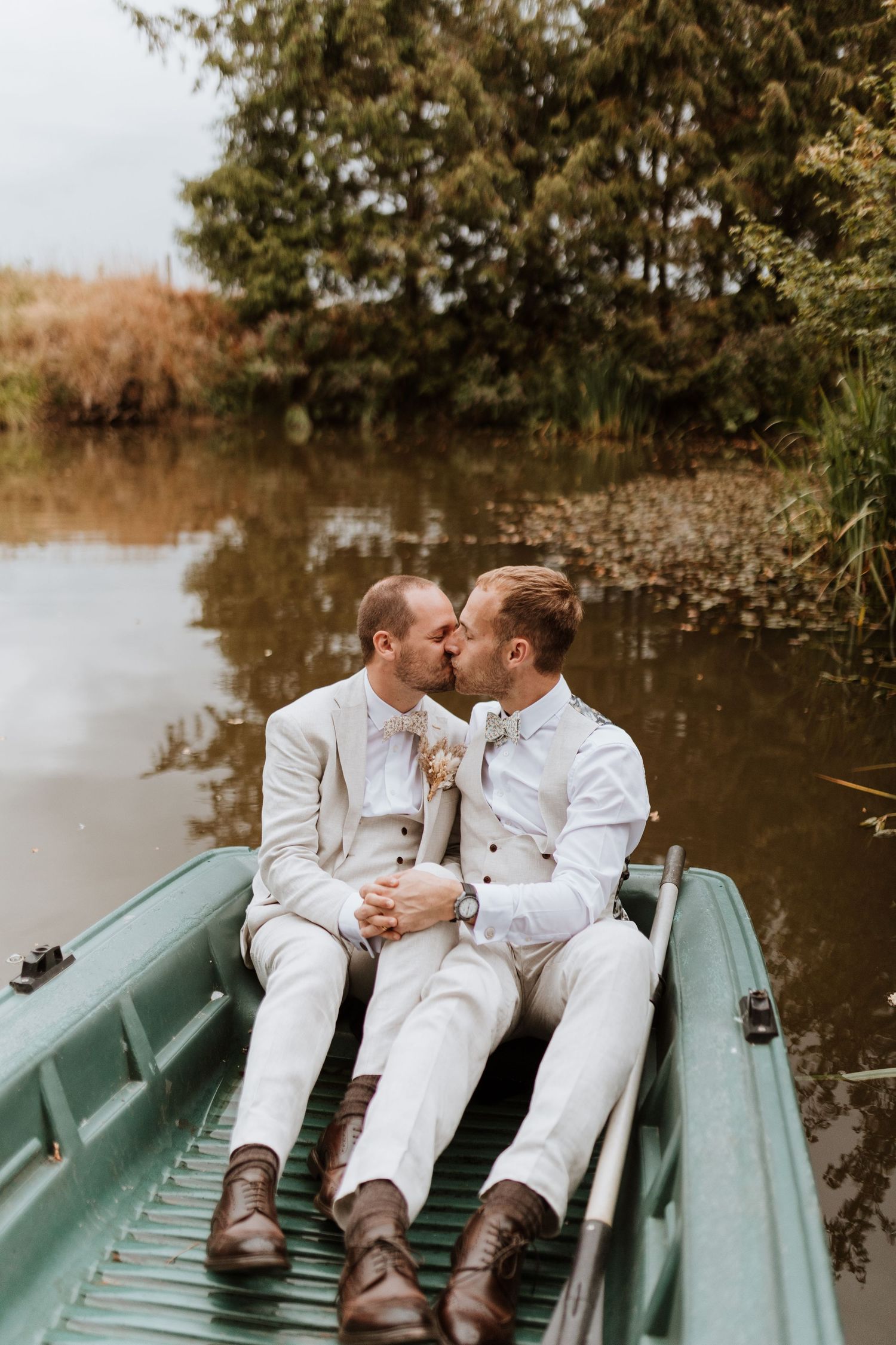 Two grooms in white suits share a romantic kiss while sitting in a green rowboat on a calm lake surrounded by trees at Moat Hall Barns