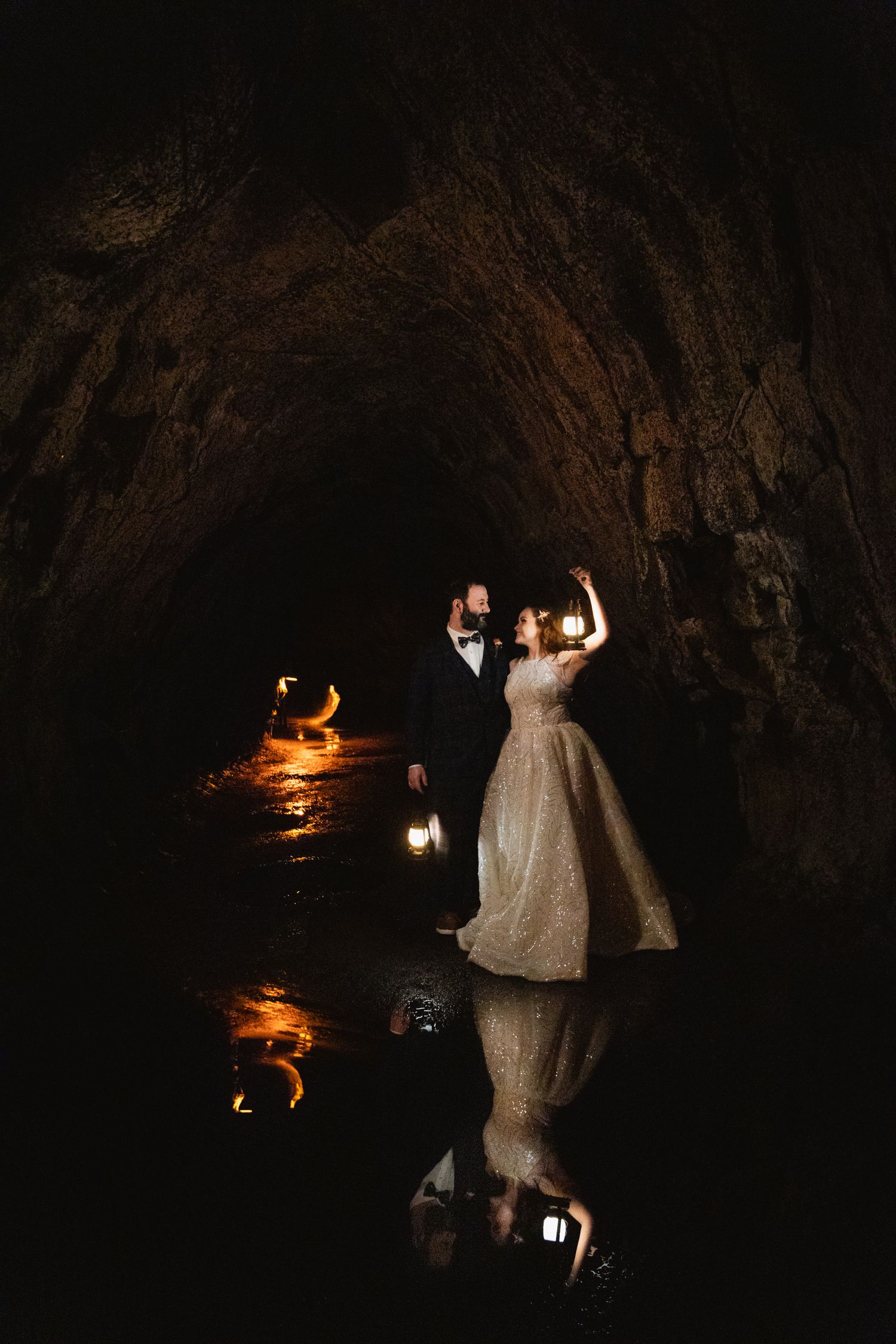 Bride and groom holding lanterns in a dark cave with glowing water reflections.