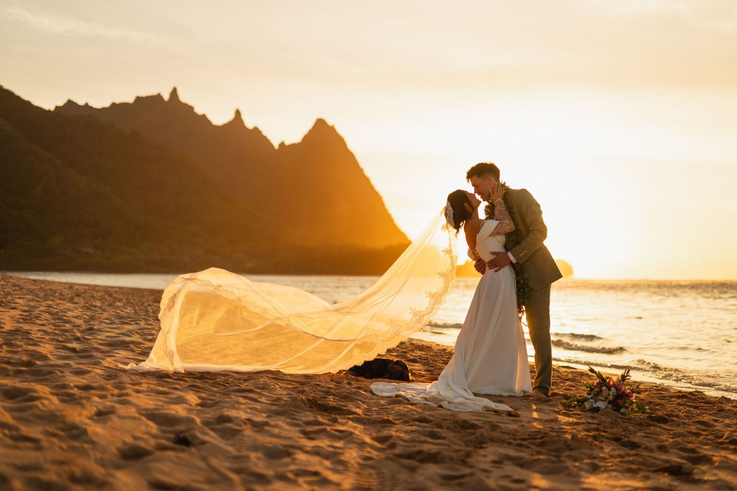 Bride and groom kiss at golden sunset on beach with veil billowing and mountains behind.