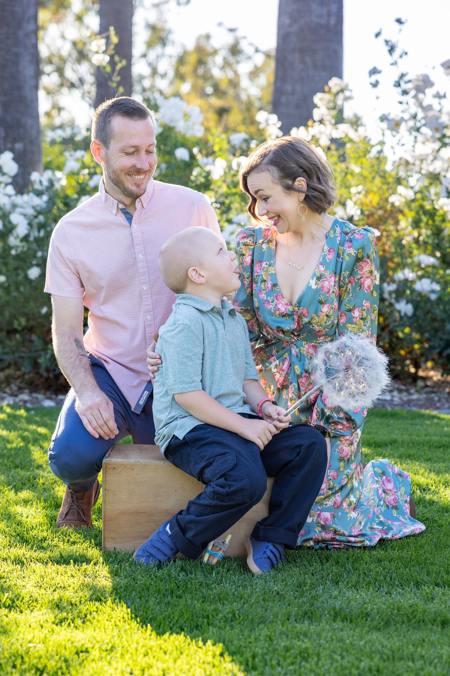 Mother in floral dress sitting on green grass with baby boy in her lap, surrounded by blooming white roses.