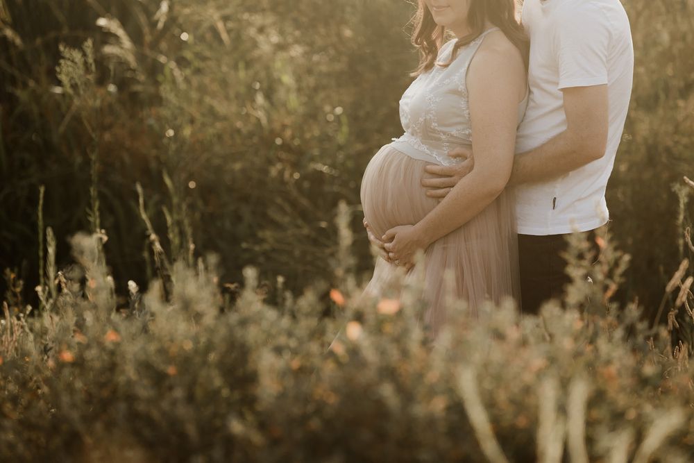 Pregnant couple embracing outdoors in golden sunlight surrounded by wildflowers, maternity photography in nature.