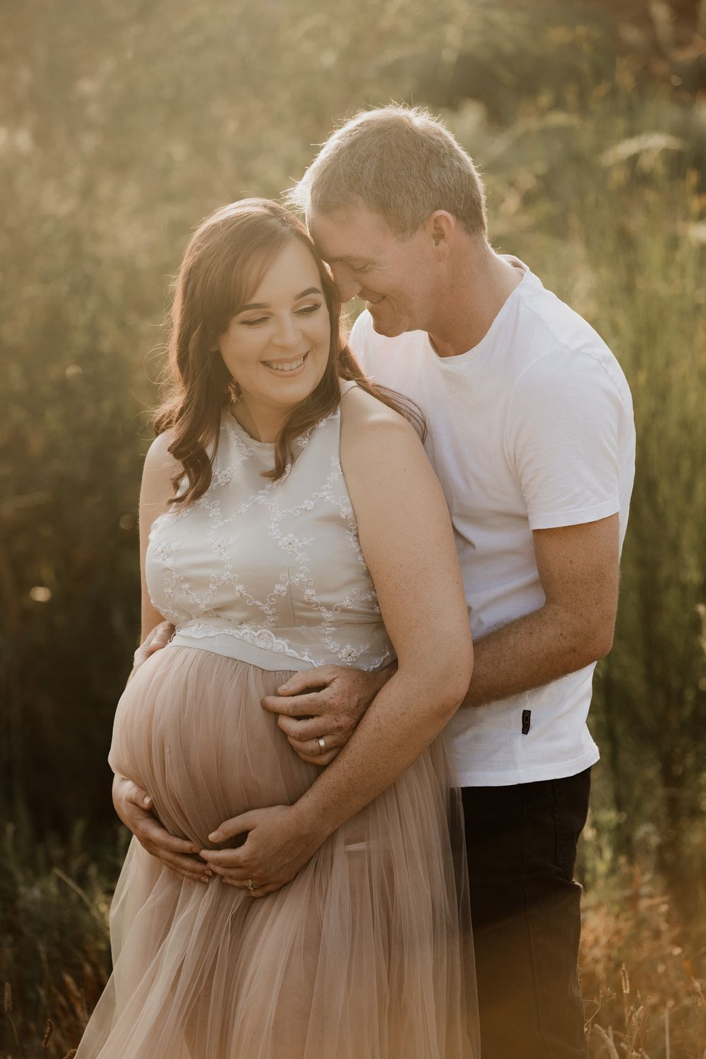 Couple sharing a tender maternity moment outdoors in golden sunlight, woman in lace top and blush tulle skirt cradling baby bump.