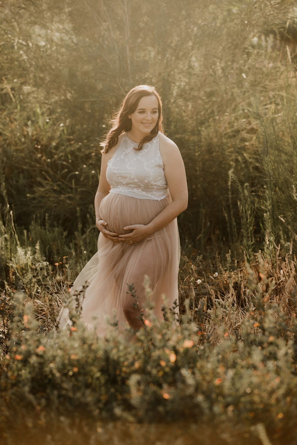 Pregnant woman in white lace top and blush tulle skirt cradling her baby bump in a sunlit field.