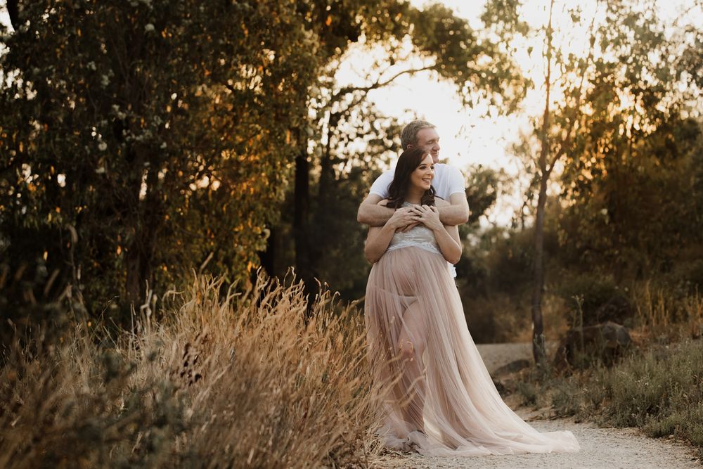 Couple embracing on a sunlit path surrounded by golden grass and trees during a romantic outdoor maternity photo session.