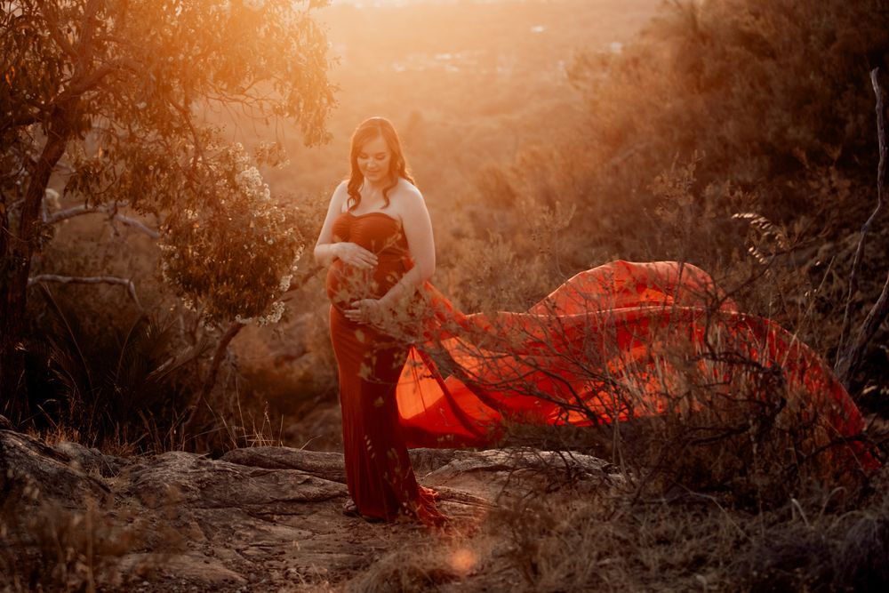 Pregnant woman in flowing red skirt and white top poses on rocks at golden sunset in natural outdoor setting.