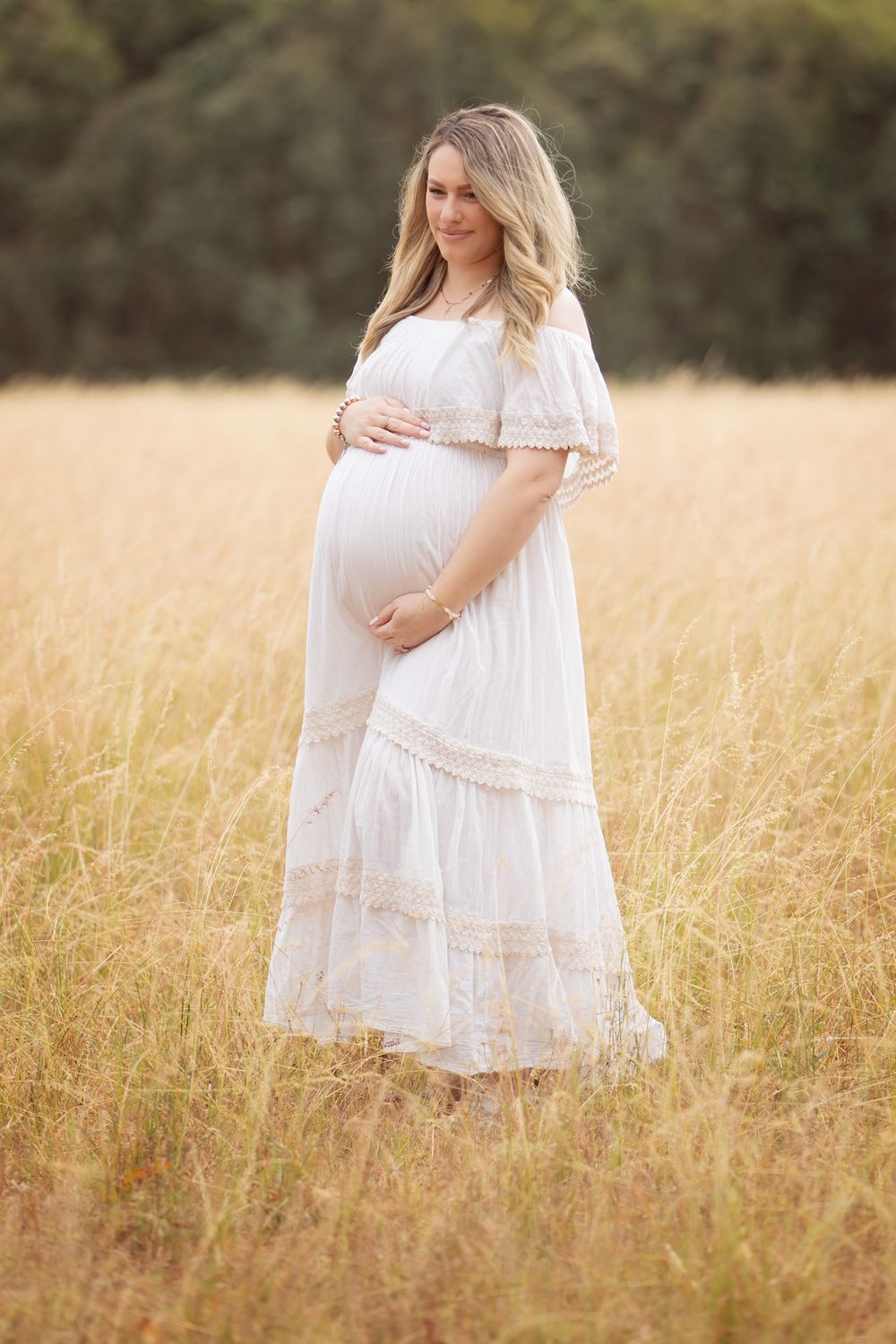 Pregnant woman in white boho dress standing in golden field, cradling baby bump, with lush green trees in background.