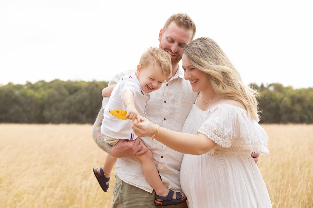 Happy family with toddler boy standing in golden wheat field, mother in white dress, father holding child with yellow toy.