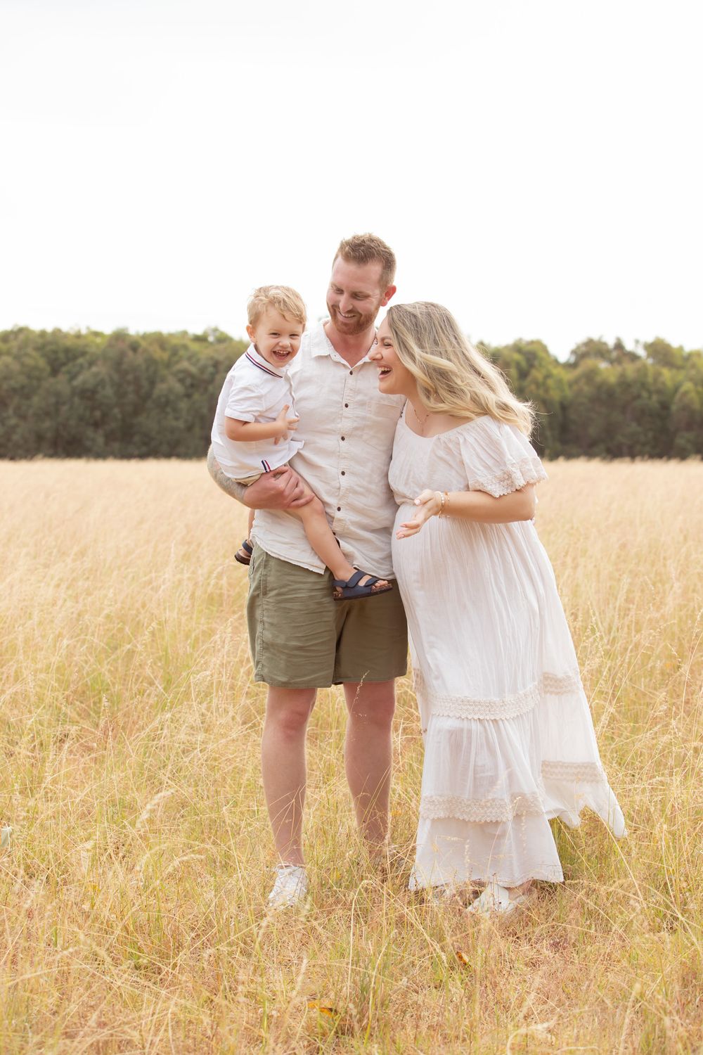 Pregnant woman in white dress with husband and toddler son standing in golden grass field for maternity photo shoot.