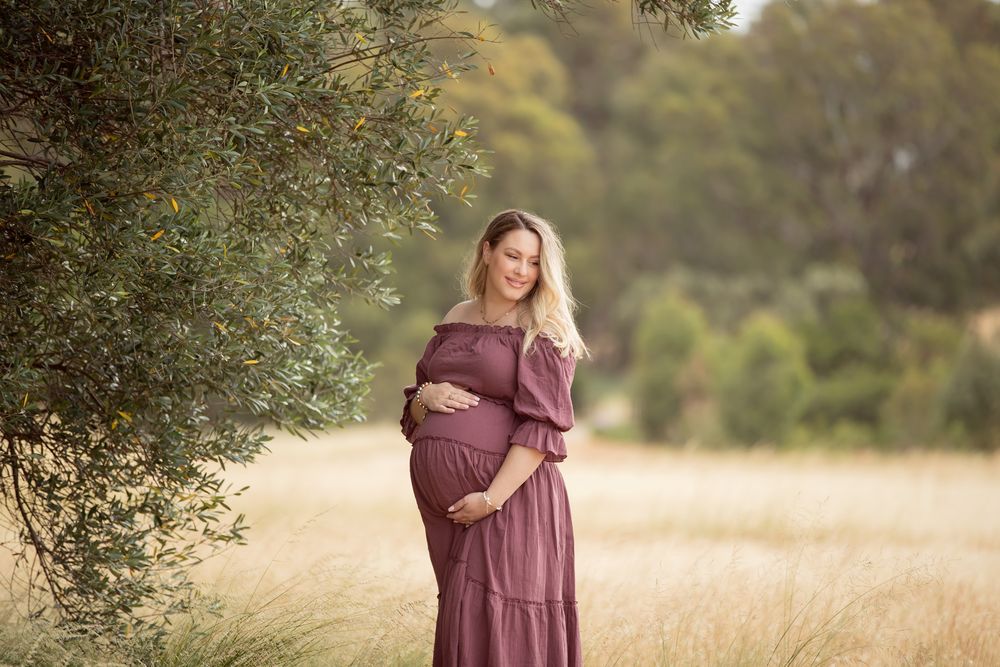 Pregnant woman in mauve off-shoulder dress cradling baby bump in golden field near olive tree.