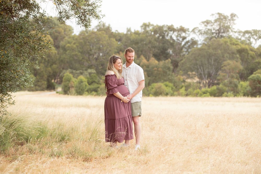 Couple standing in golden field, woman in burgundy maternity dress, man in white shirt, surrounded by trees.
