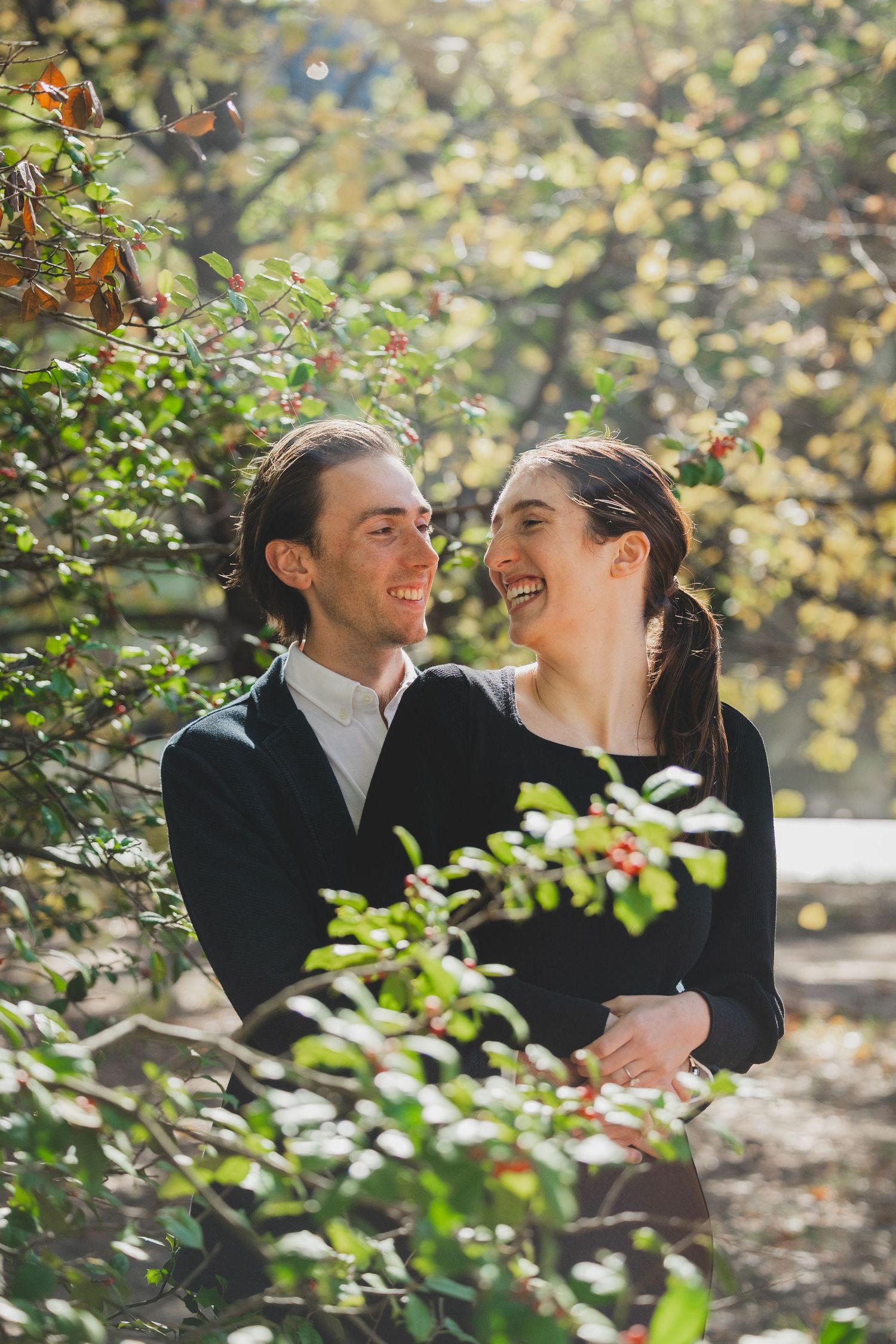 A smiling couple in black attire shares a romantic moment surrounded by lush green foliage in a sunlit park.
