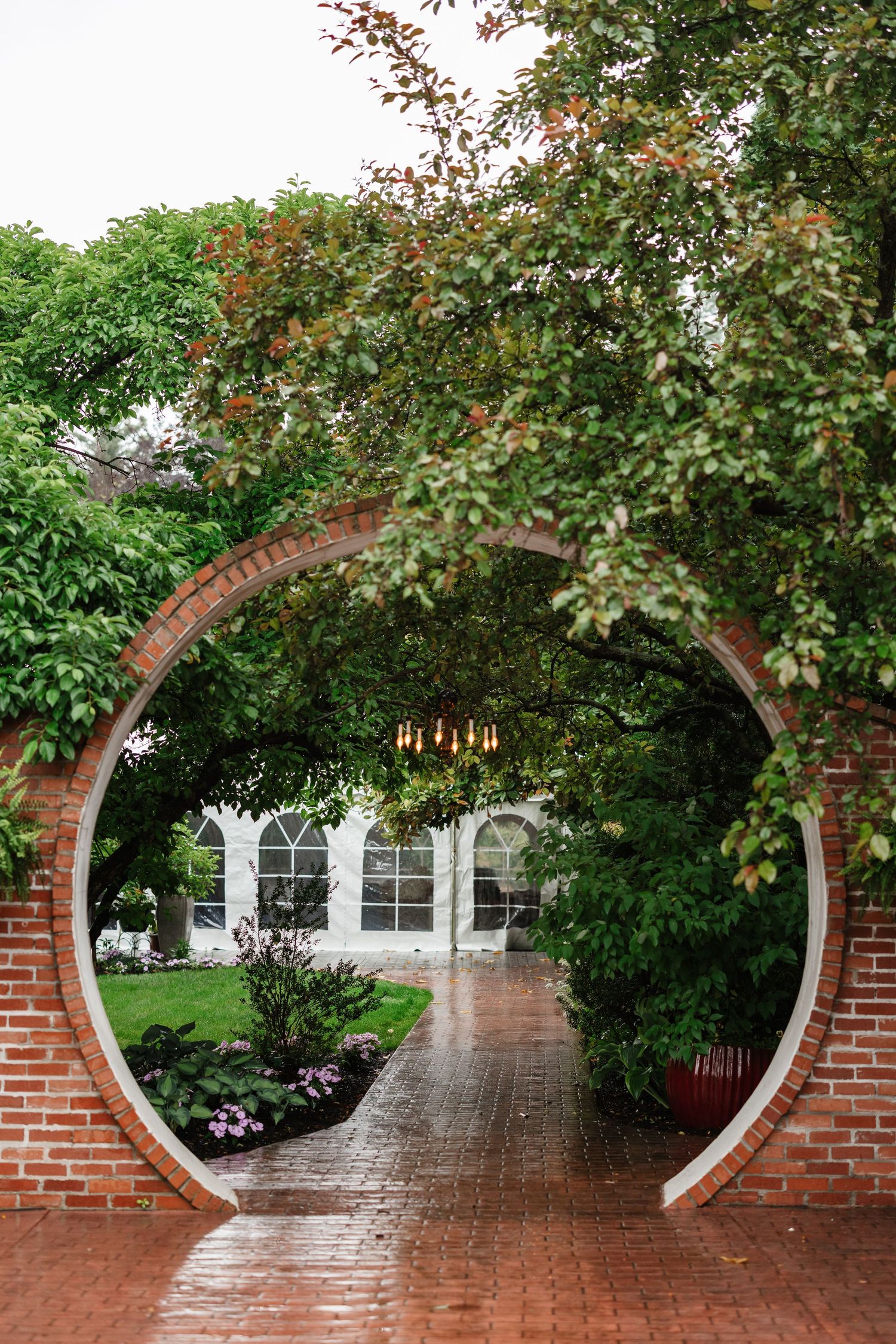 A circular brick moon gate arch framed by lush green trees leads to a white building on a rainy day.