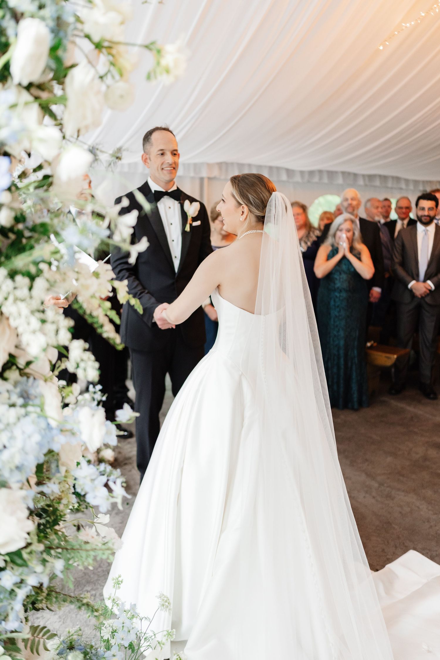 Bride in strapless white ballgown faces smiling groom at floral arch during elegant tent wedding ceremony.