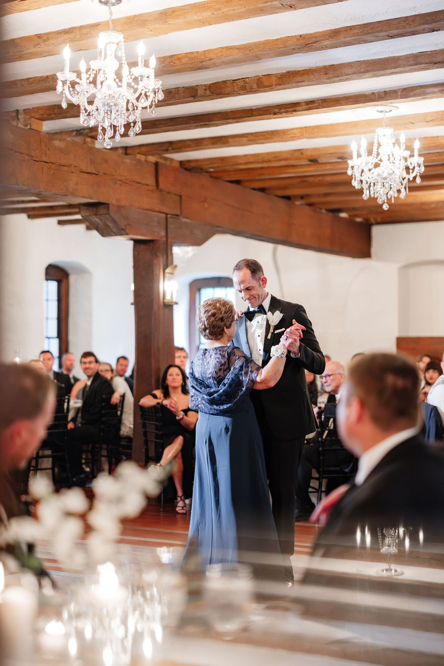 Groom dances with mother in blue gown under crystal chandeliers at wedding reception.