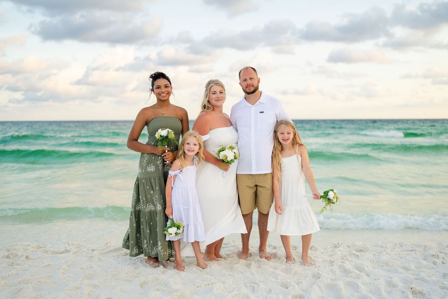 Beach wedding party with bride, groom, bridesmaid, and two flower girls posing on white sand with turquoise ocean backdrop.