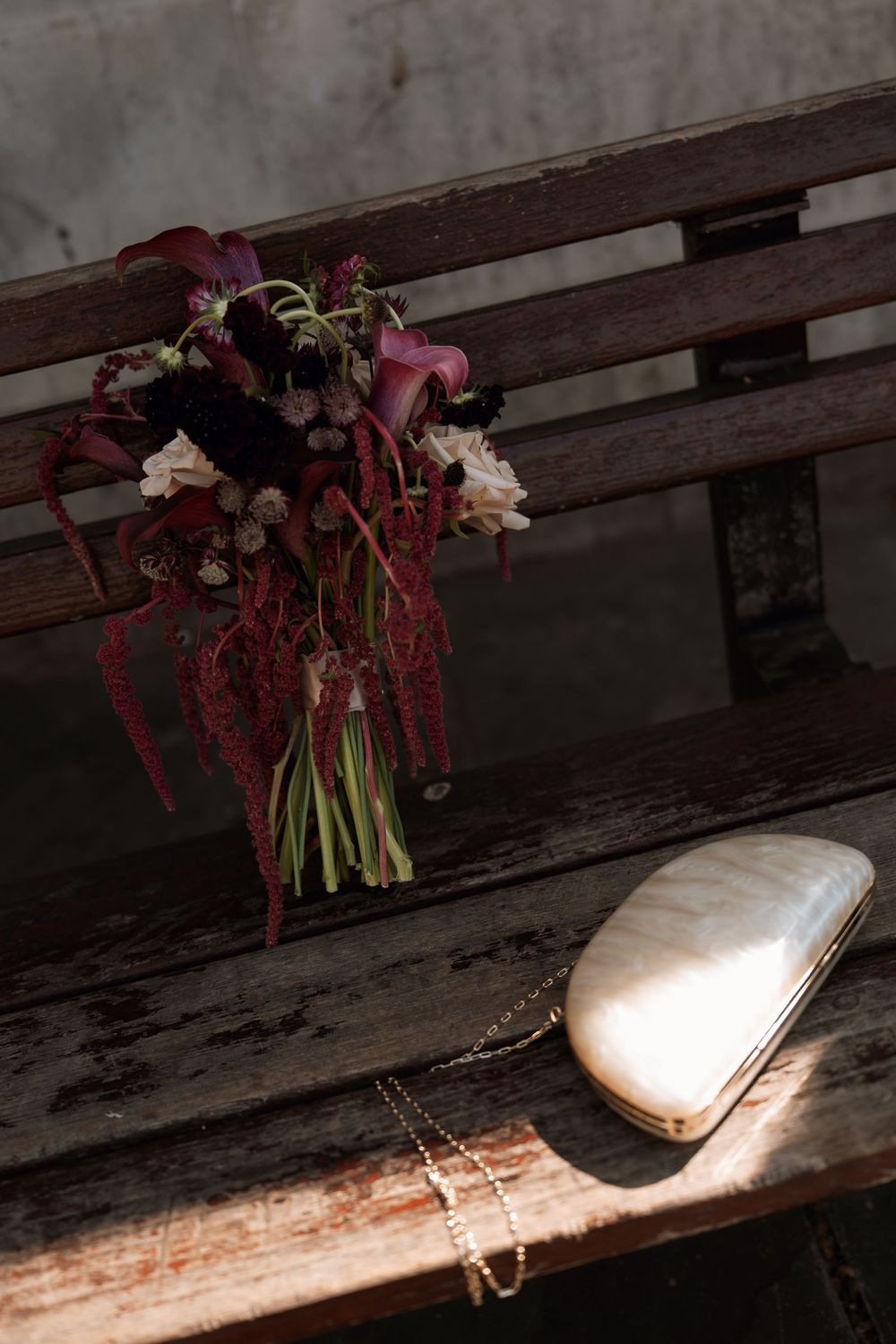 Dark floral bouquet with burgundy amaranthus and calla lilies beside a silver clutch purse on a rustic wooden bench.