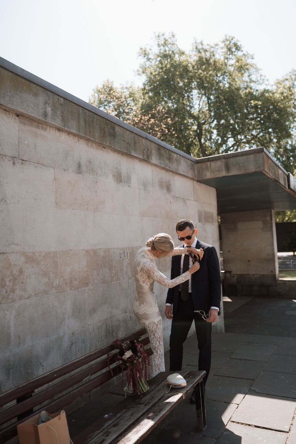 Bride adjusting groom's tie outdoors near stone wall and bench on sunny wedding day.