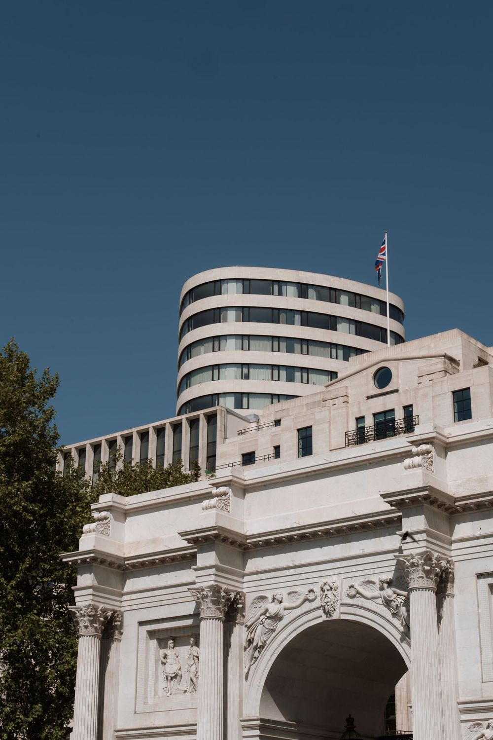 White marble arch with ornate carvings stands before a curved modern building flying a British flag under clear blue sky.