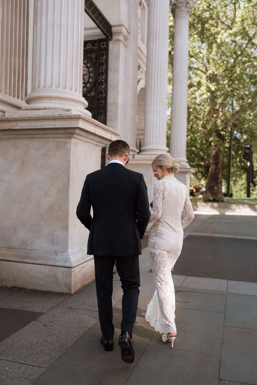 Bride in lace gown and groom in black suit walk past white marble columns at elegant wedding venue.