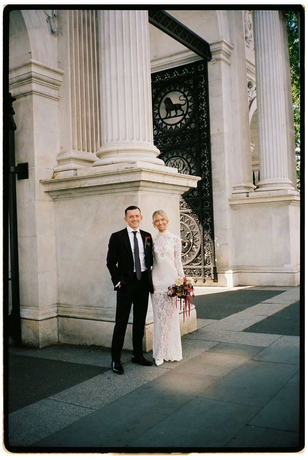 A bride and groom pose in wedding attire before grand stone columns and ornate iron gates.