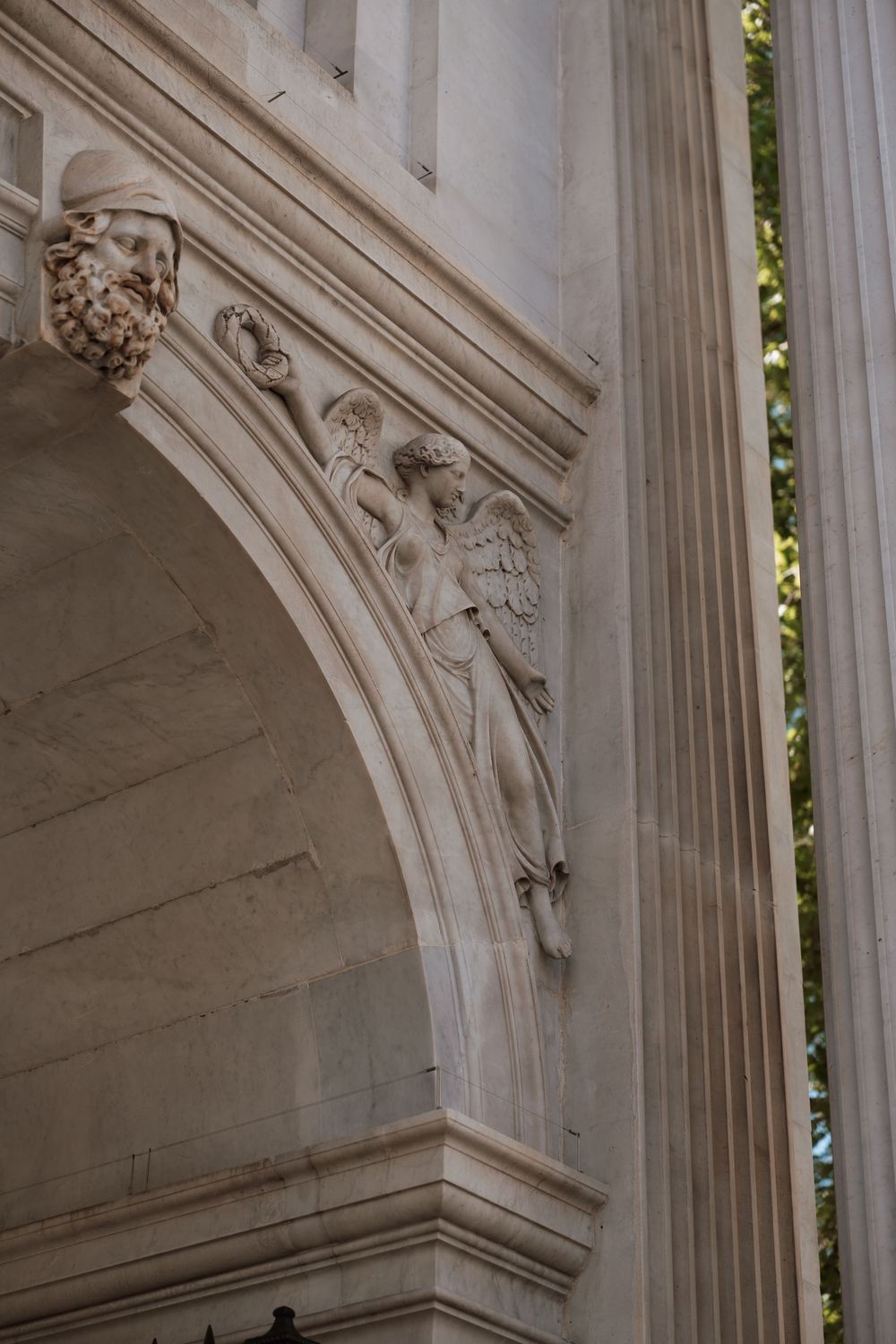Classical stone arch with carved angel sculpture and decorative keystones on a neoclassical building facade.