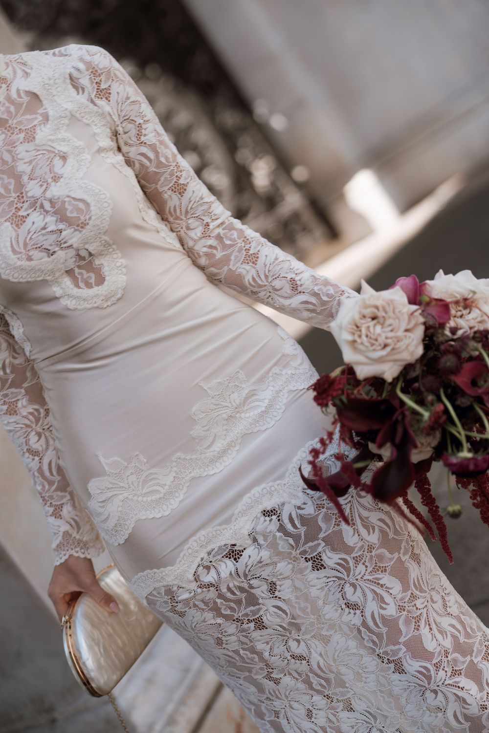 Pregnant bride in white lace long-sleeve gown holding burgundy bouquet and ivory clutch.