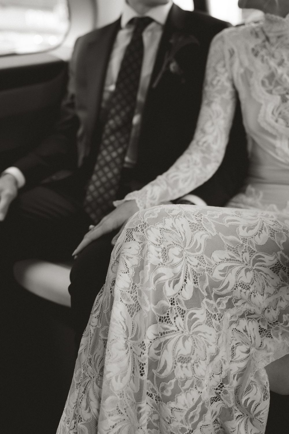 Black and white photo of bride in lace gown and groom in suit sitting together in a car on their wedding day.
