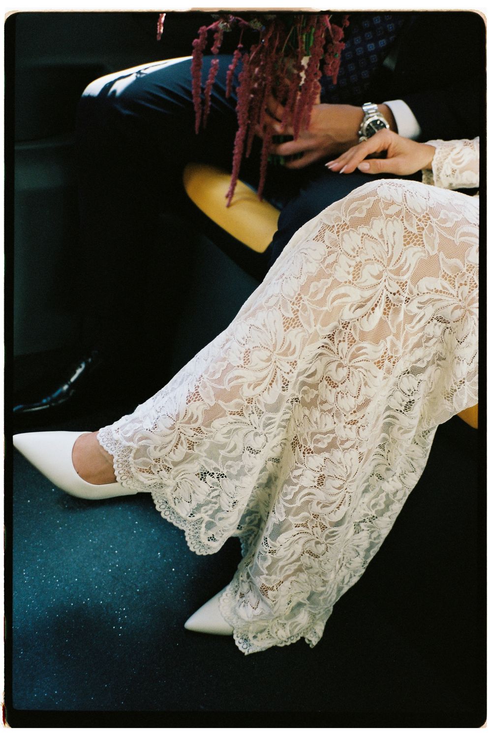 Bride in white lace dress and pointed white heels seated beside groom in dark suit holding pink flowers.