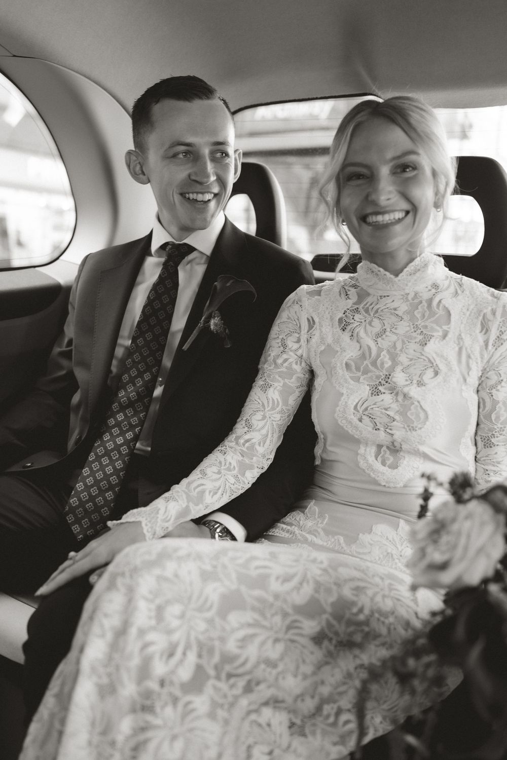 Black and white photo of a smiling bride and groom seated in a car on their wedding day.