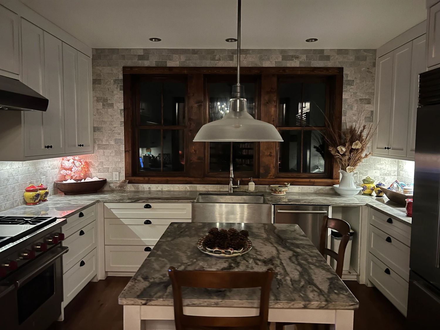 Modern kitchen with white cabinets, granite island, pendant light, stone backsplash, and stainless steel appliances at night.