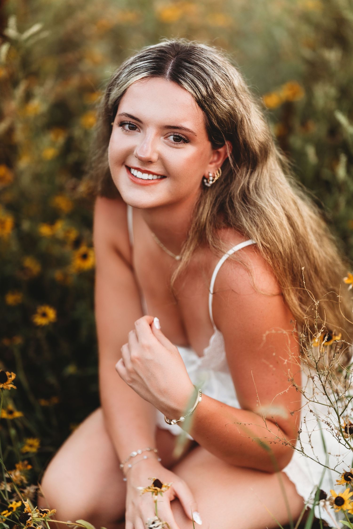 Young woman with long blonde hair smiles in a sunflower field, wearing a white dress and delicate jewelry.