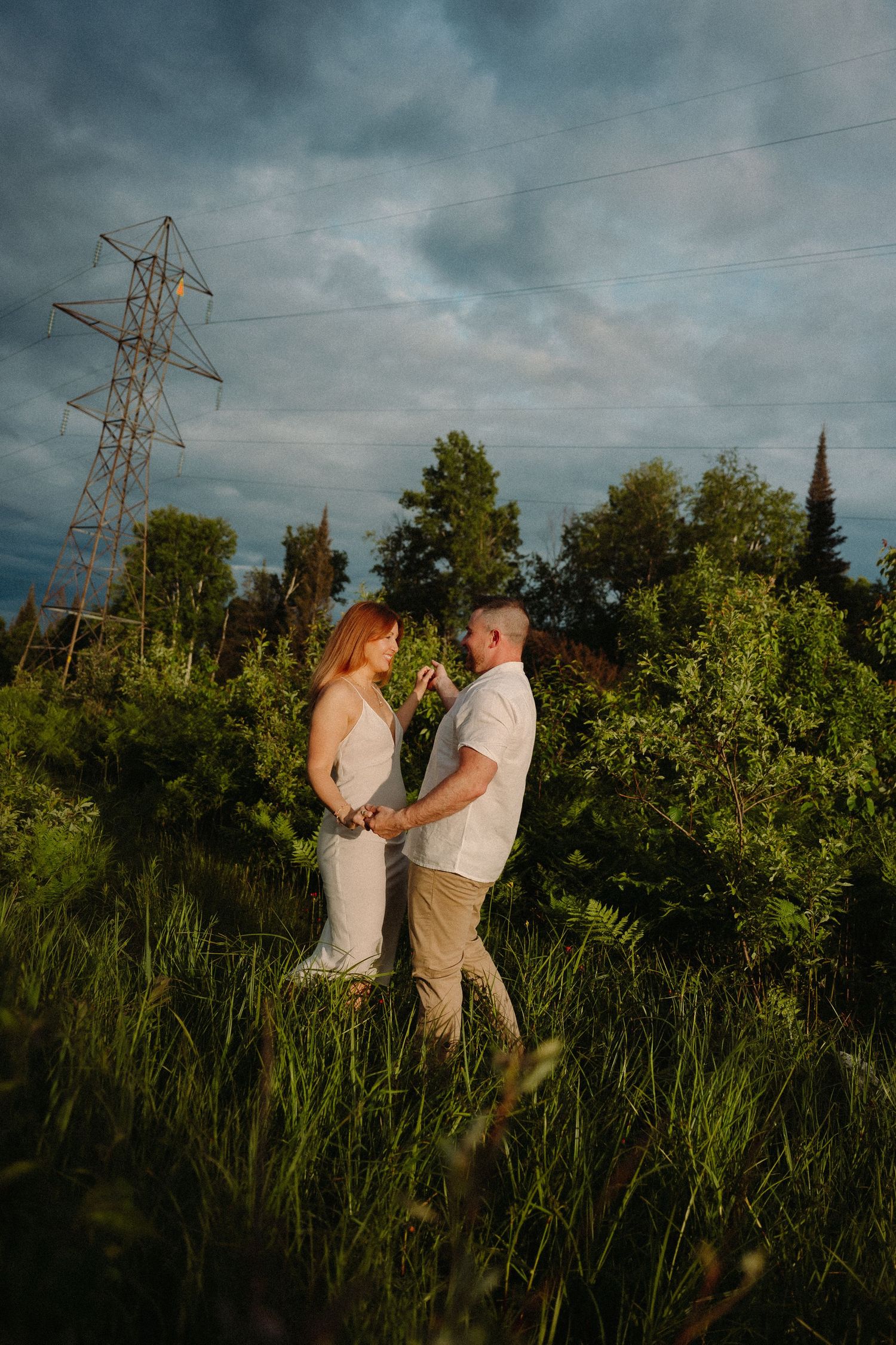 Couple holds hands in lush green field near power tower under dramatic cloudy sky at golden hour.