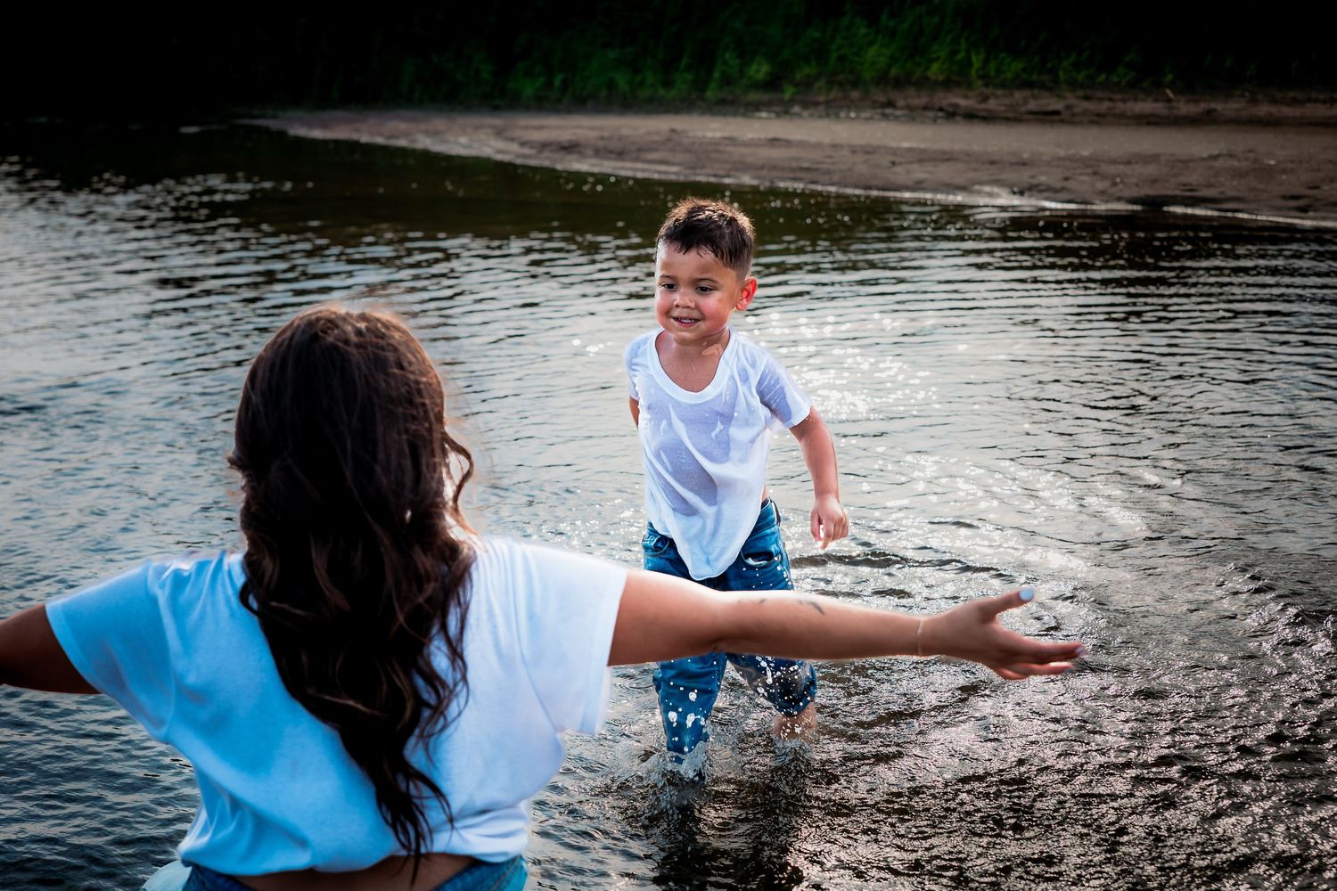 A young boy splashes in a shallow lake toward a woman with outstretched arms on a summer day.