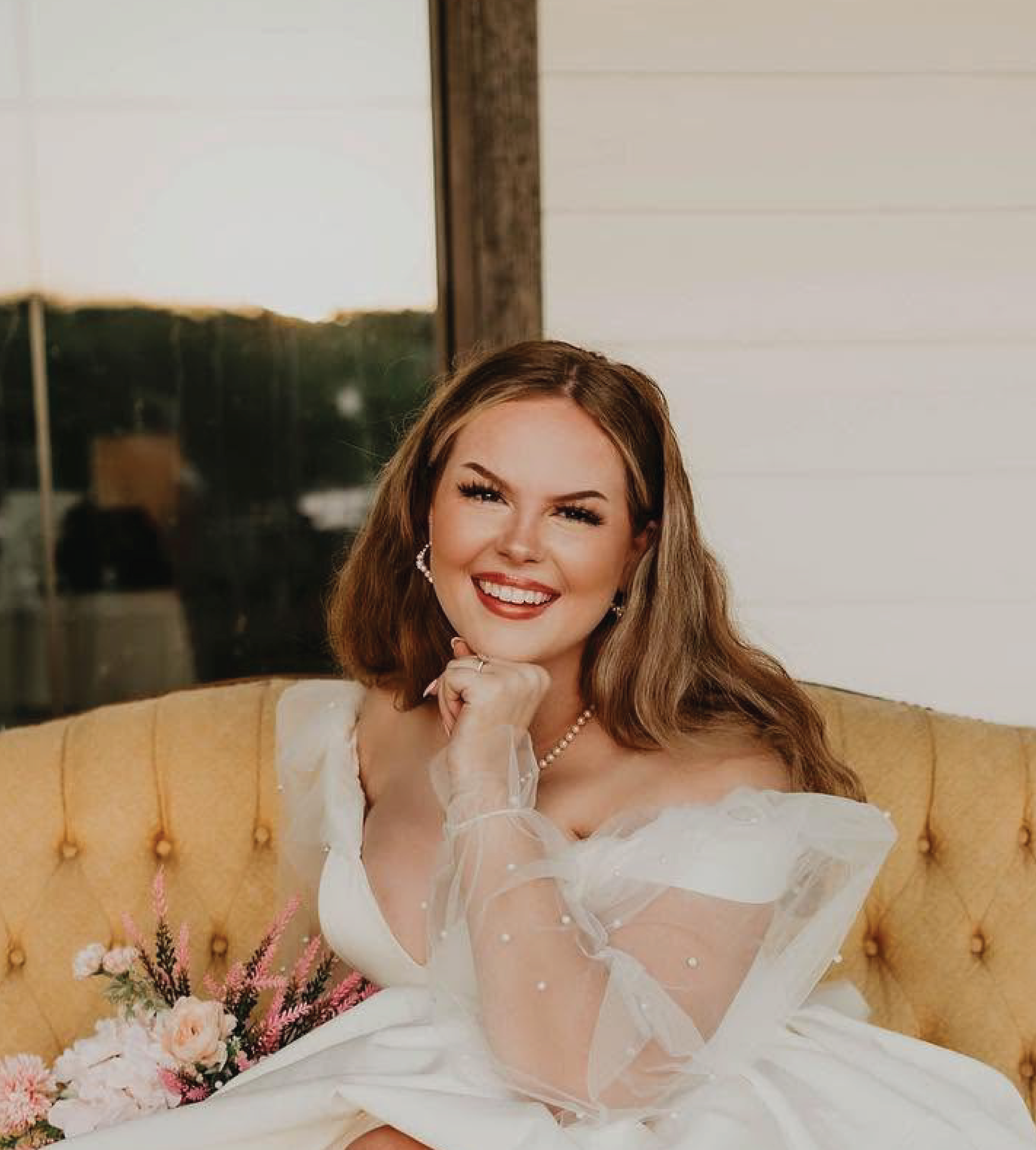 Smiling bride in a white off-shoulder gown, pearl necklace, seated on a gold velvet sofa, holding a pink floral. About the photographer.