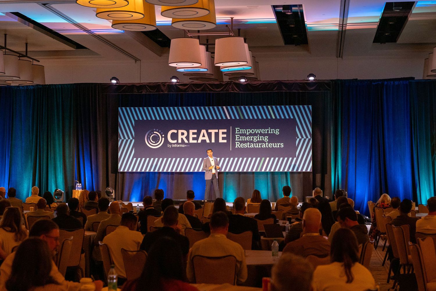 Speaker presenting at a formal conference event with an attentive audience seated in a darkened banquet hall.