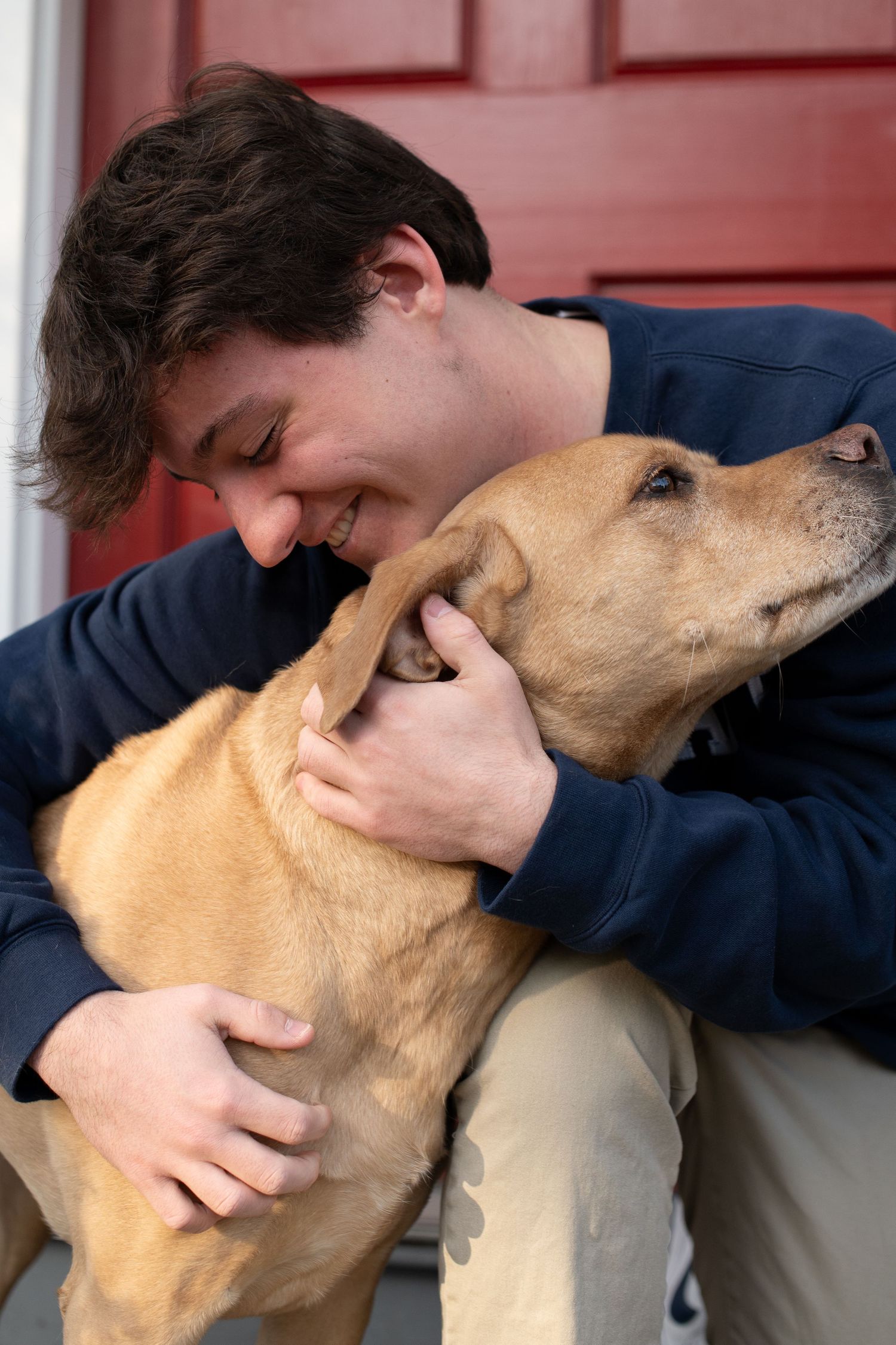 A young man in a navy sweatshirt hugs and nuzzles a tan Labrador dog on front steps by a red door.