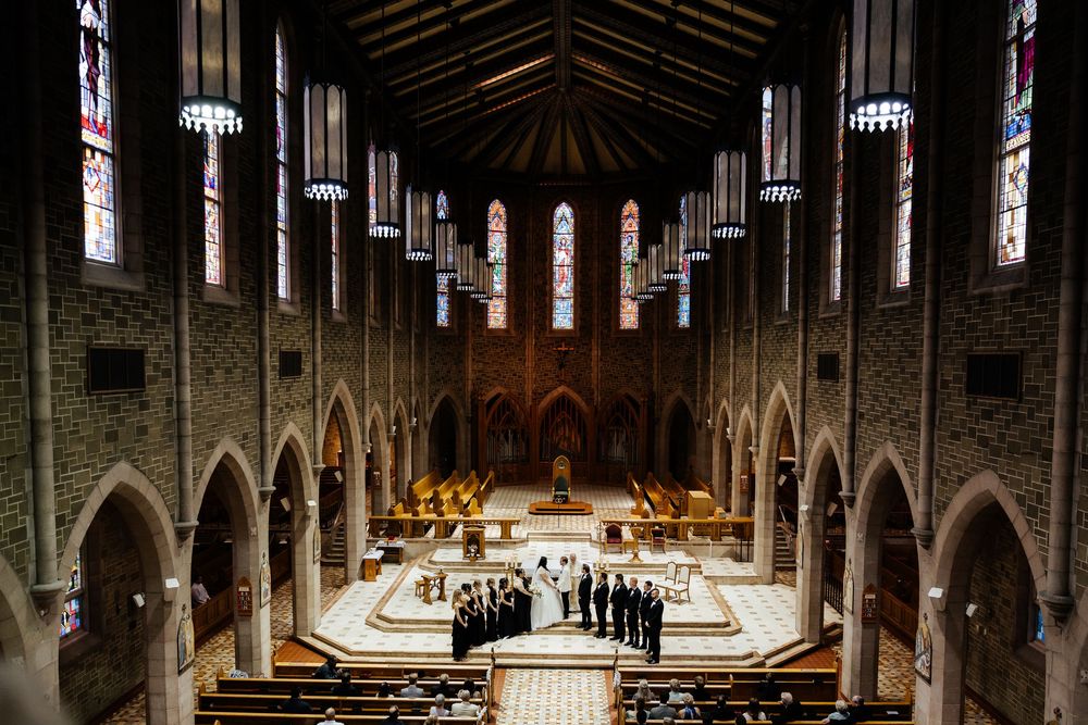 Grand Gothic church interior with choir performing at altar, featuring arched stone walls and vibrant stained glass windows.