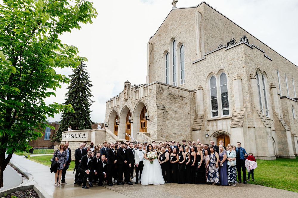 Wedding party poses outside a grand stone basilica church surrounded by lush green trees on a cloudy day.