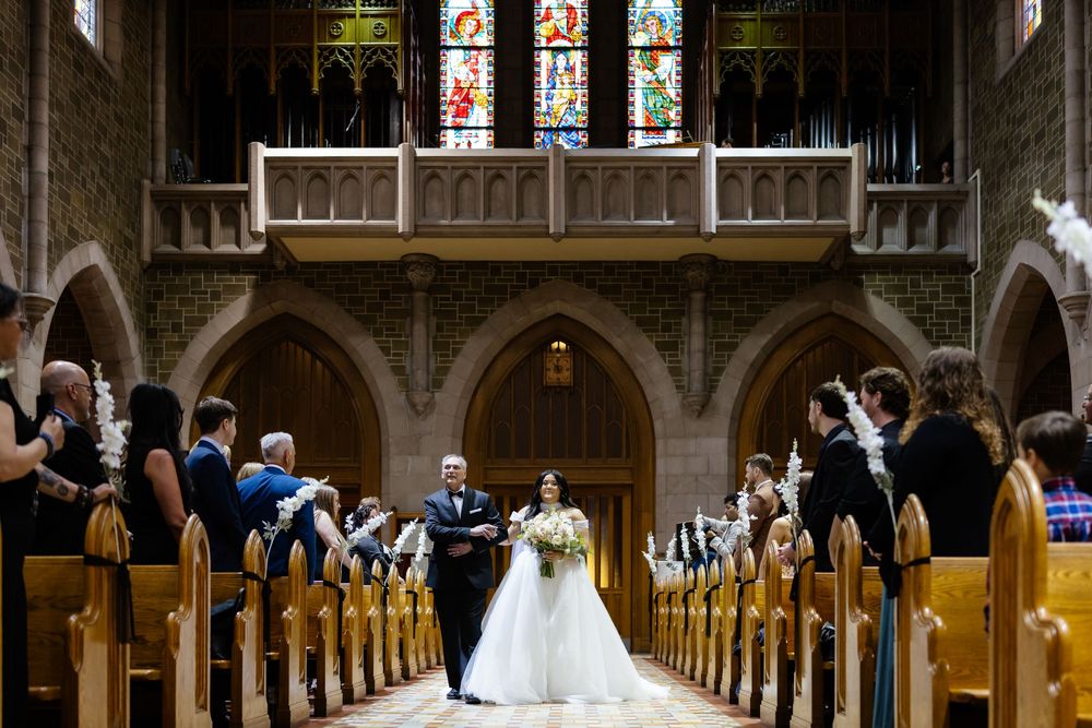 Bride walks down church aisle with escort past wooden pews and stained glass windows during wedding ceremony.