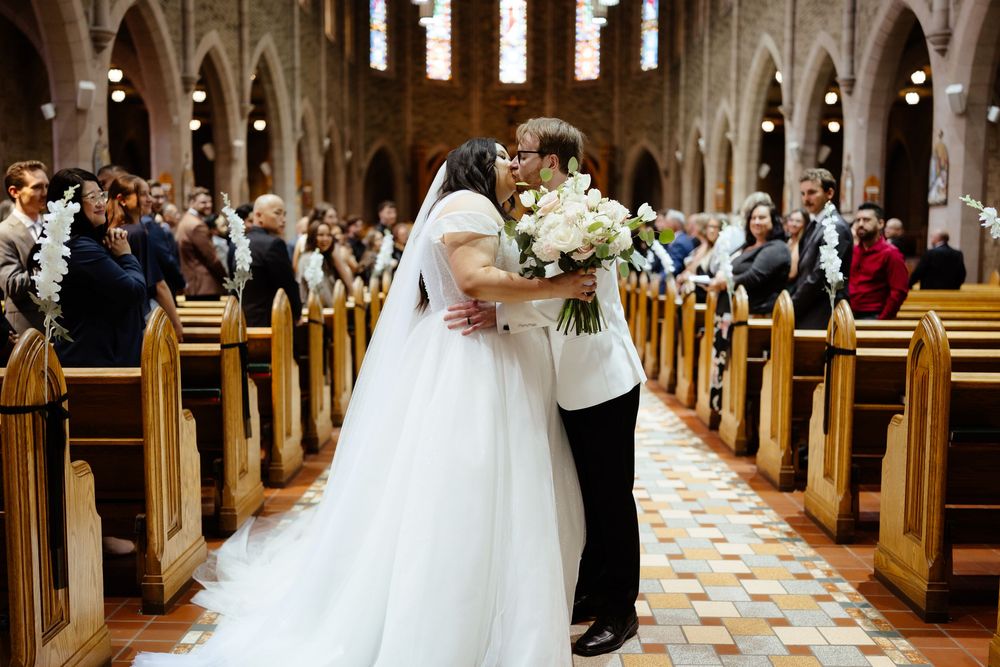 Bride and groom share first kiss in elegant church aisle, bride holding white bouquet, guests seated in wooden pews.