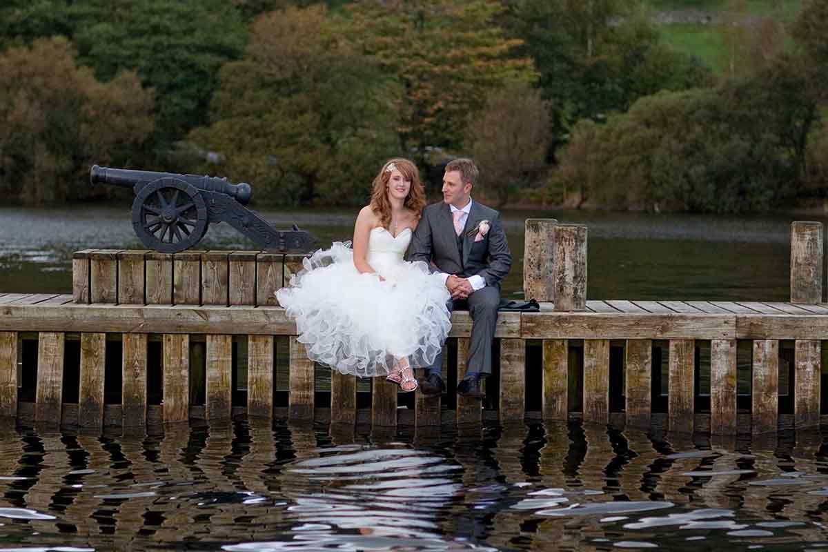 Bride and groom sitting on a wooden pier by a lake with a vintage cannon in the background.