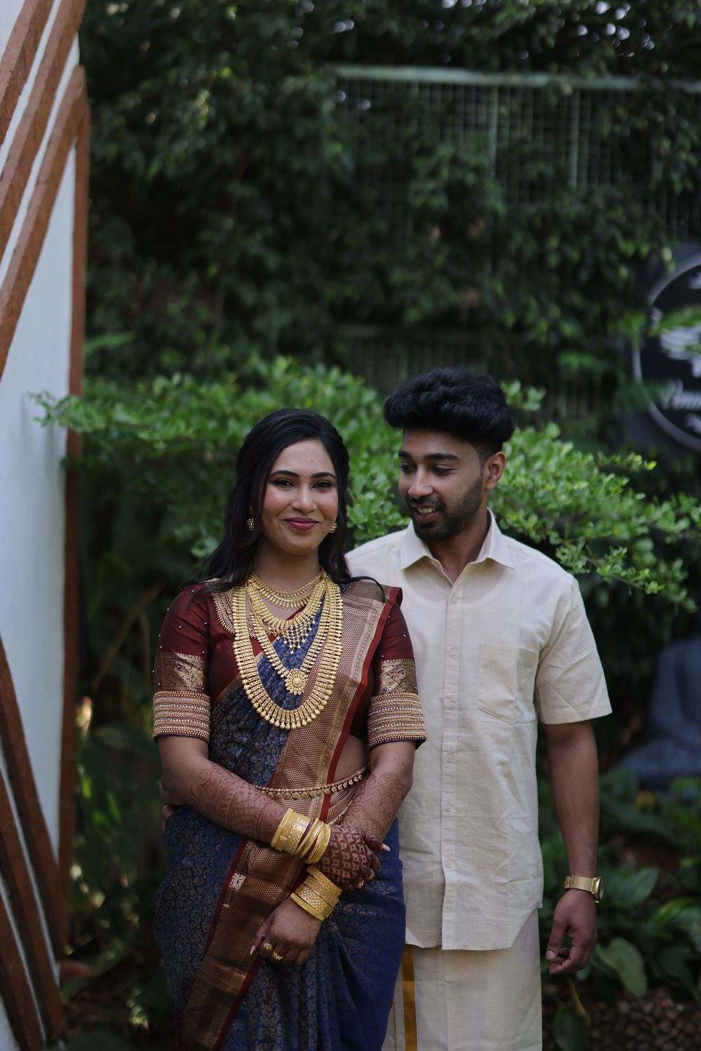 South Indian couple in traditional attire, woman in silk saree with gold jewelry, man in cream shirt and dhoti, posing outdoors.