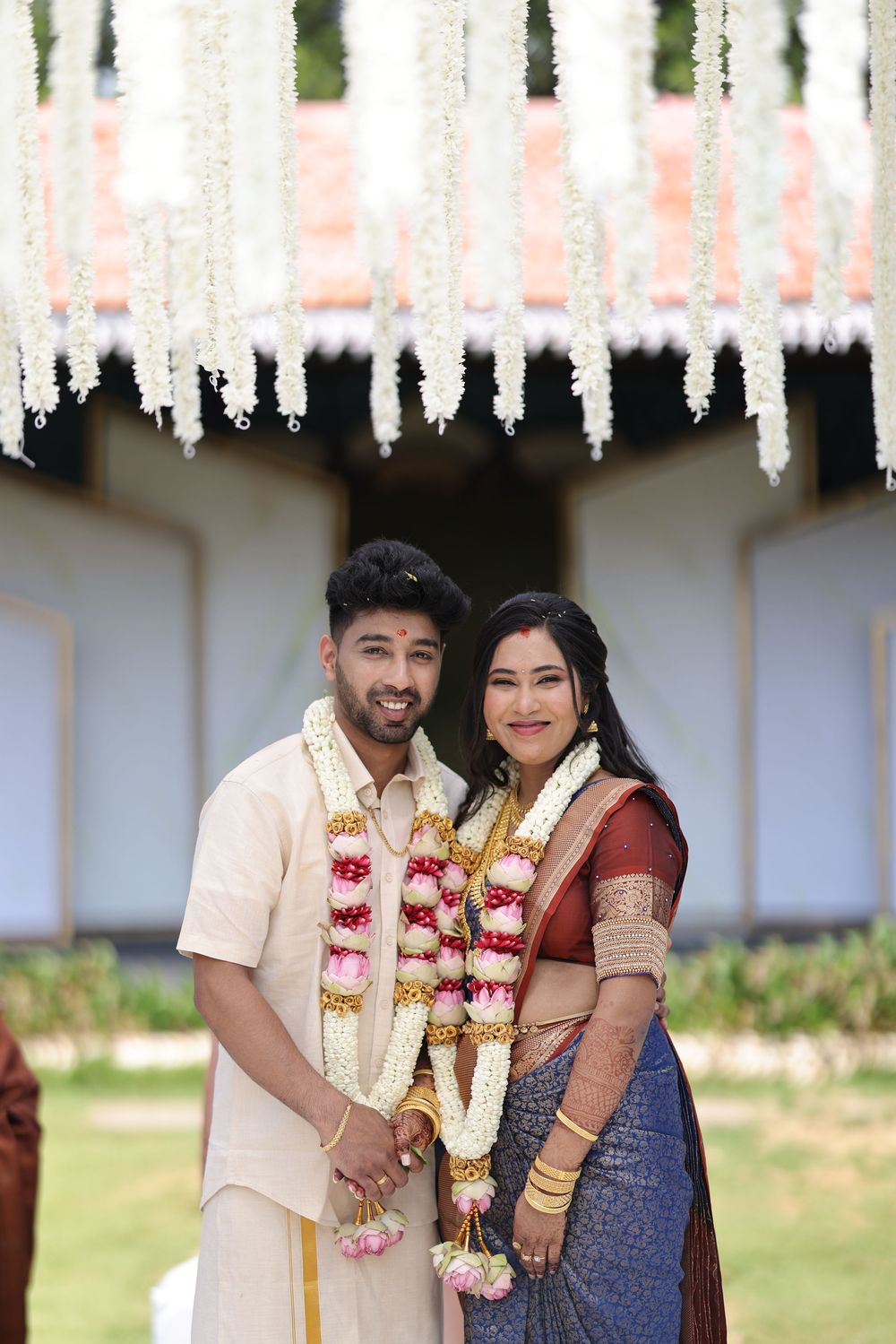 Couple in traditional South Indian wedding attire with floral garlands posing under white flower decorations.