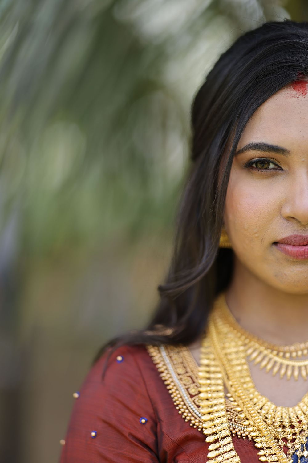 Young Indian woman in traditional red outfit with gold jewelry and red bindi, posing outdoors near palm trees.