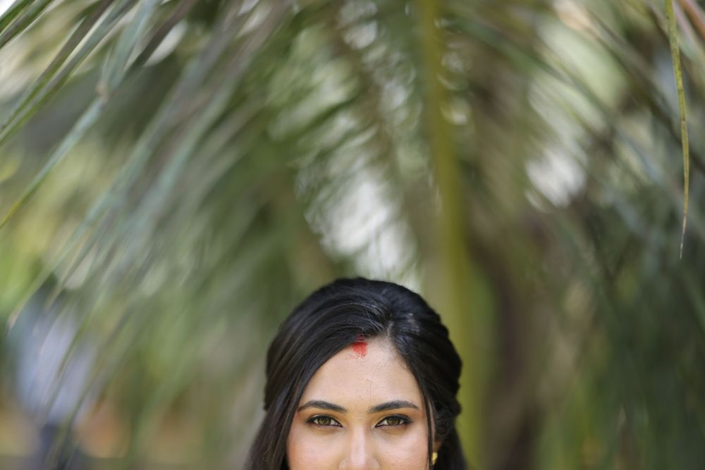 A young Indian woman with a red bindi stands gracefully beneath lush green palm fronds outdoors.