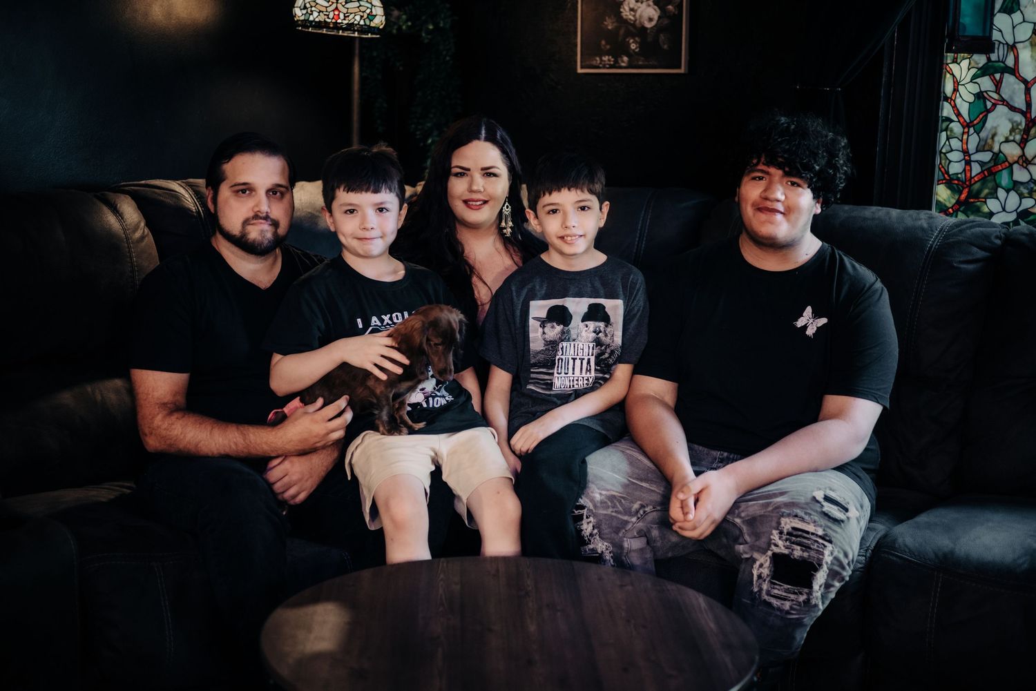 Family of five posing together on a dark sofa, smiling warmly in a cozy, moody living room setting.
