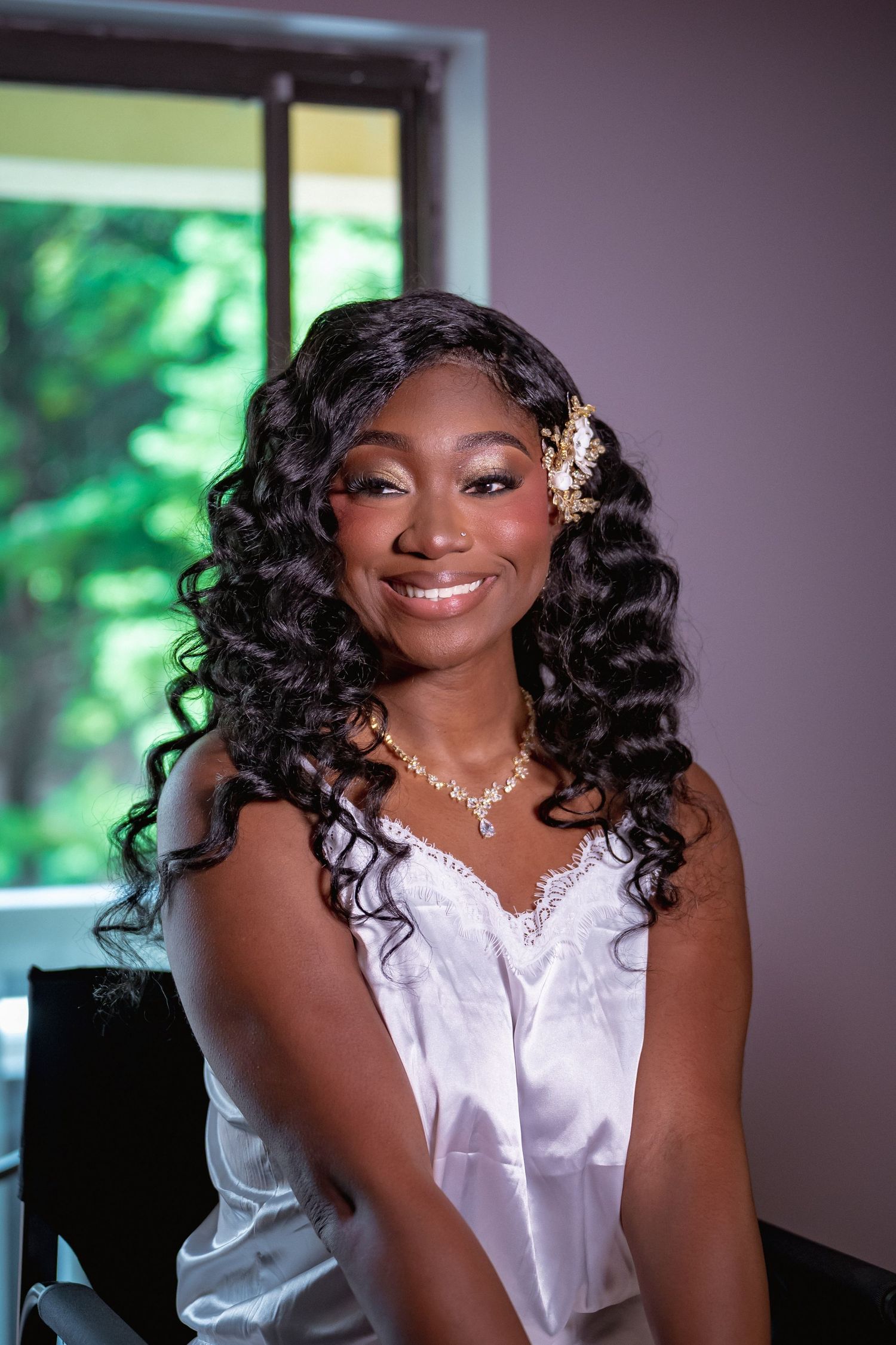 Smiling bride with curly hair, gold hair accessory, white dress, and pearl necklace sitting near a window.