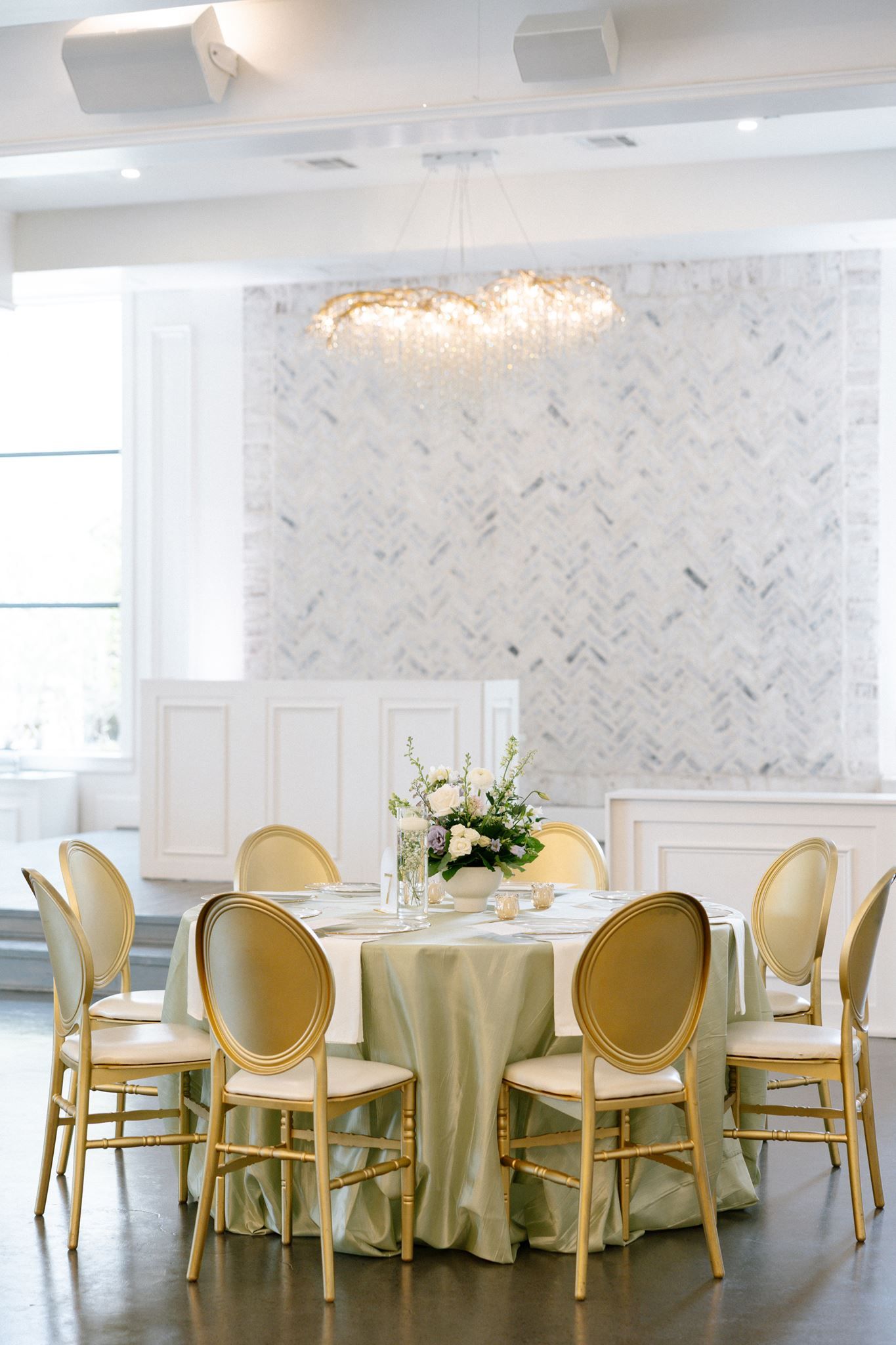 Elegant wedding reception table with gold chairs, sage green linen, floral centerpiece, and crystal chandelier in a bright white venue.